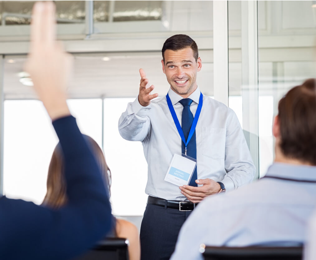 A man in a tie and blue lanyard around his neck smiling as he calls on someone raising their hand in a meeting